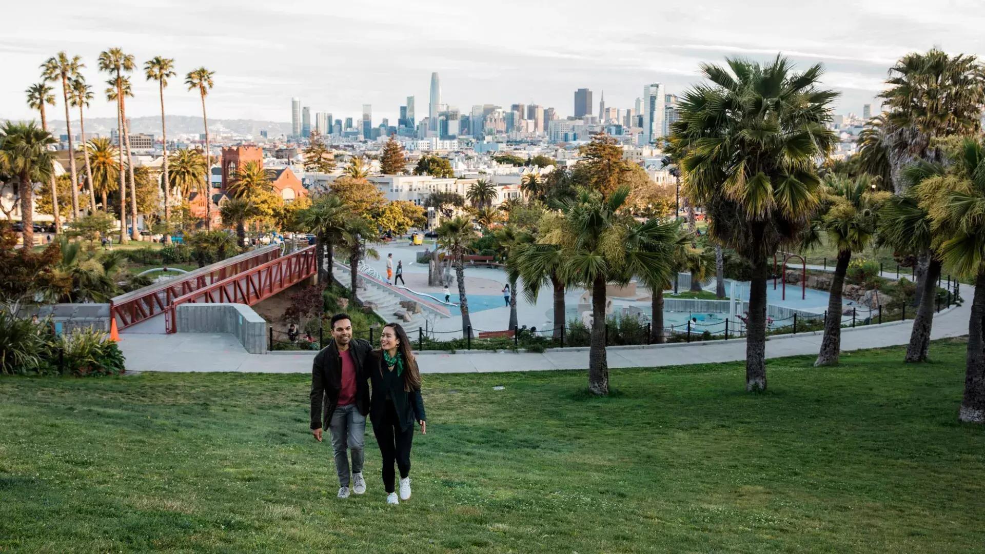 Un couple se dirige vers la caméra avec Dolores Park et la Skyline de San Francisco derrière eux.