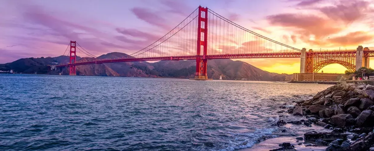的 Golden Gate Bridge at sunset with a multicolored sky and the San Francisco Bay in the foreground.